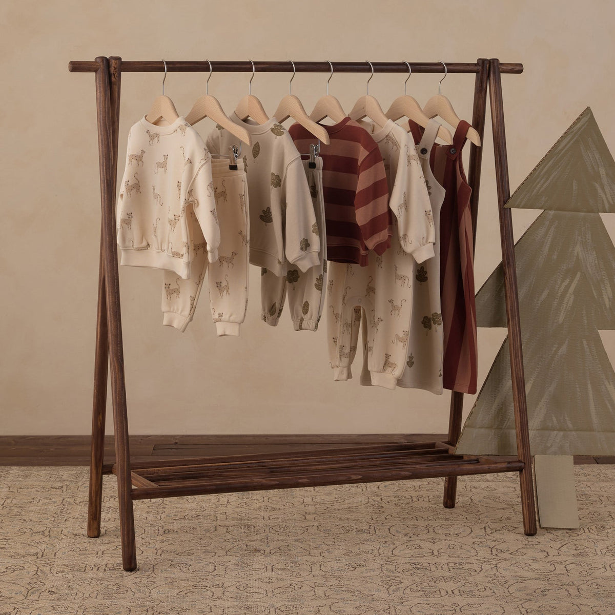 A wooden A-frame clothing rack holding several children's long-sleeve tops and shorts in cream, beige, and burgundy tones, styled in a warm neutral room with a patterned rug and a decorative tree on the right.