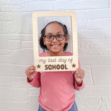 Child holding a sign saying 'my last day of school' against a white brick wall.