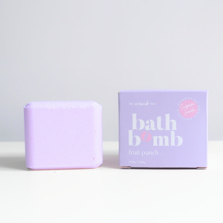 A lavender bath bomb next to its matching purple packaging box on a clean white surface against a light gray background. The box features the brand name 'an scrubs in' and the product name 'bath bomb fruit punch'.