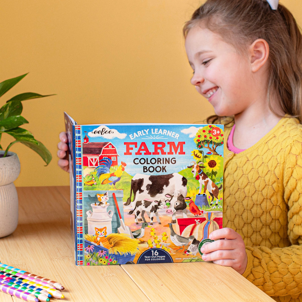 Child holding a 'Farm Coloring Book' with colorful illustrations of farm animals on a wooden table.
