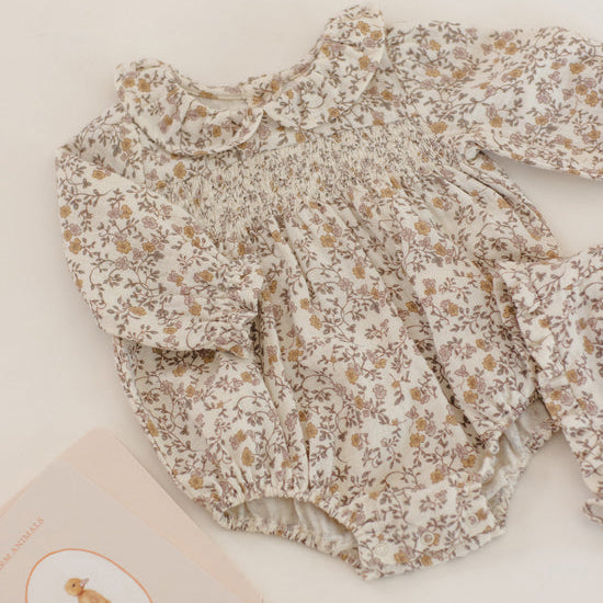 A baby long-sleeve romper with a smocked chest and ruffled collar, featuring a delicate beige and brown floral print on a cream background, styled flat-lay on a light cream surface next to a book.