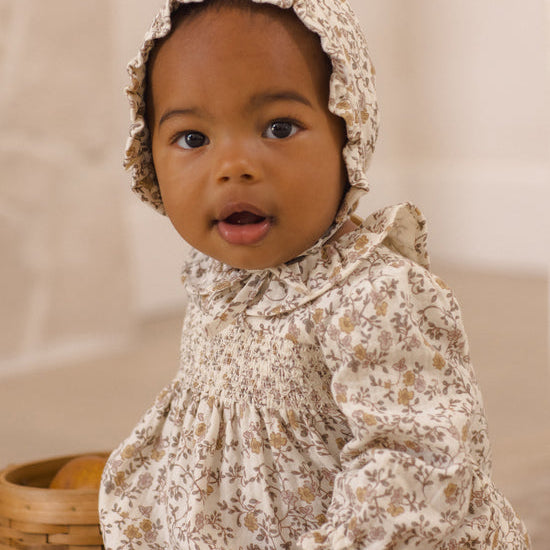 A baby wearing a cream-colored floral smocked romper and matching bonnet, sitting next to a woven basket.