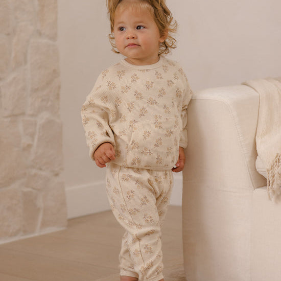 A toddler stands barefoot indoors wearing a cream-colored two-piece pajama set with a small beige floral pattern. The set includes a long-sleeve top and matching pants.
