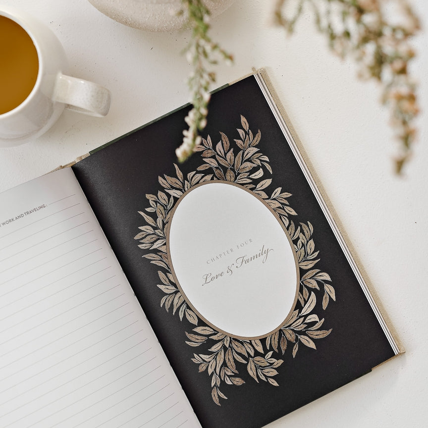 Open book with decorative cover on a white surface with a cup of coffee and flowers.