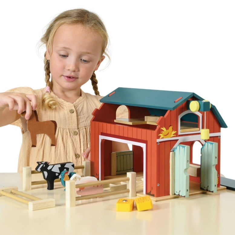 A young girl with blonde braids, wearing a short-sleeved yellow dress, plays with the Wooden Big Red Barn Playset by Mentari, holding a wooden toy horse near the open barn doors and fenced enclosure.