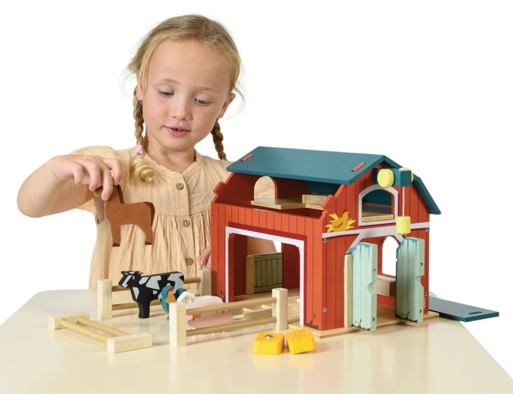 A young girl with blonde braids, wearing a short-sleeved yellow dress, plays with the Wooden Big Red Barn Playset by Mentari, holding a wooden toy horse near the open barn doors and fenced enclosure.