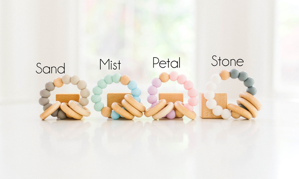 Four pairs of wooden bead teething rings labeled Sand, Mist, Petal, and Stone, arranged on a white surface with a soft blurred background.
