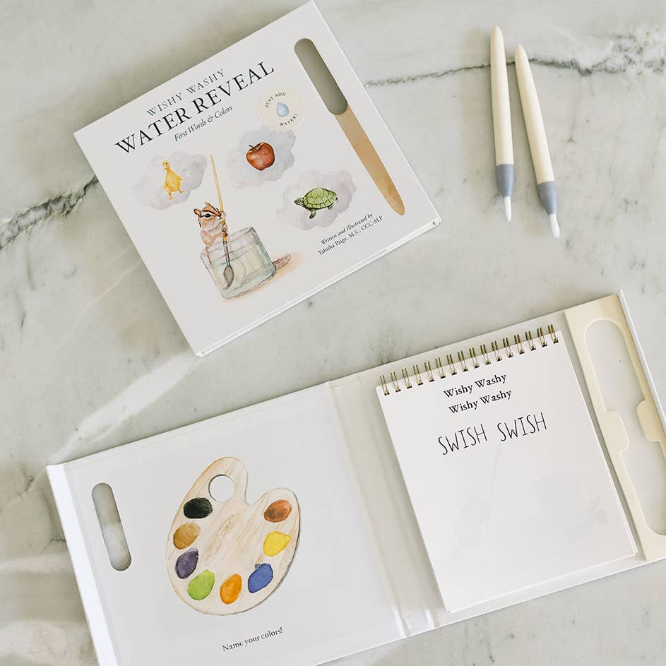 A children's water painting kit shown open on a light marble surface, with the illustrated box, a wooden palette tool, two white paint pens, and a spiral-bound notebook page that reads 'Whiny Washy Wasty Wry Wry SWISH SWISH'.