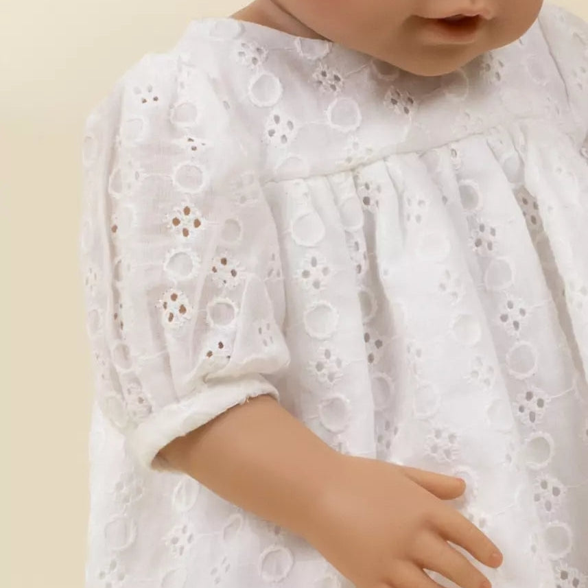 A close-up of a doll or toy baby wearing a white eyelet lace dress against a plain light beige background.