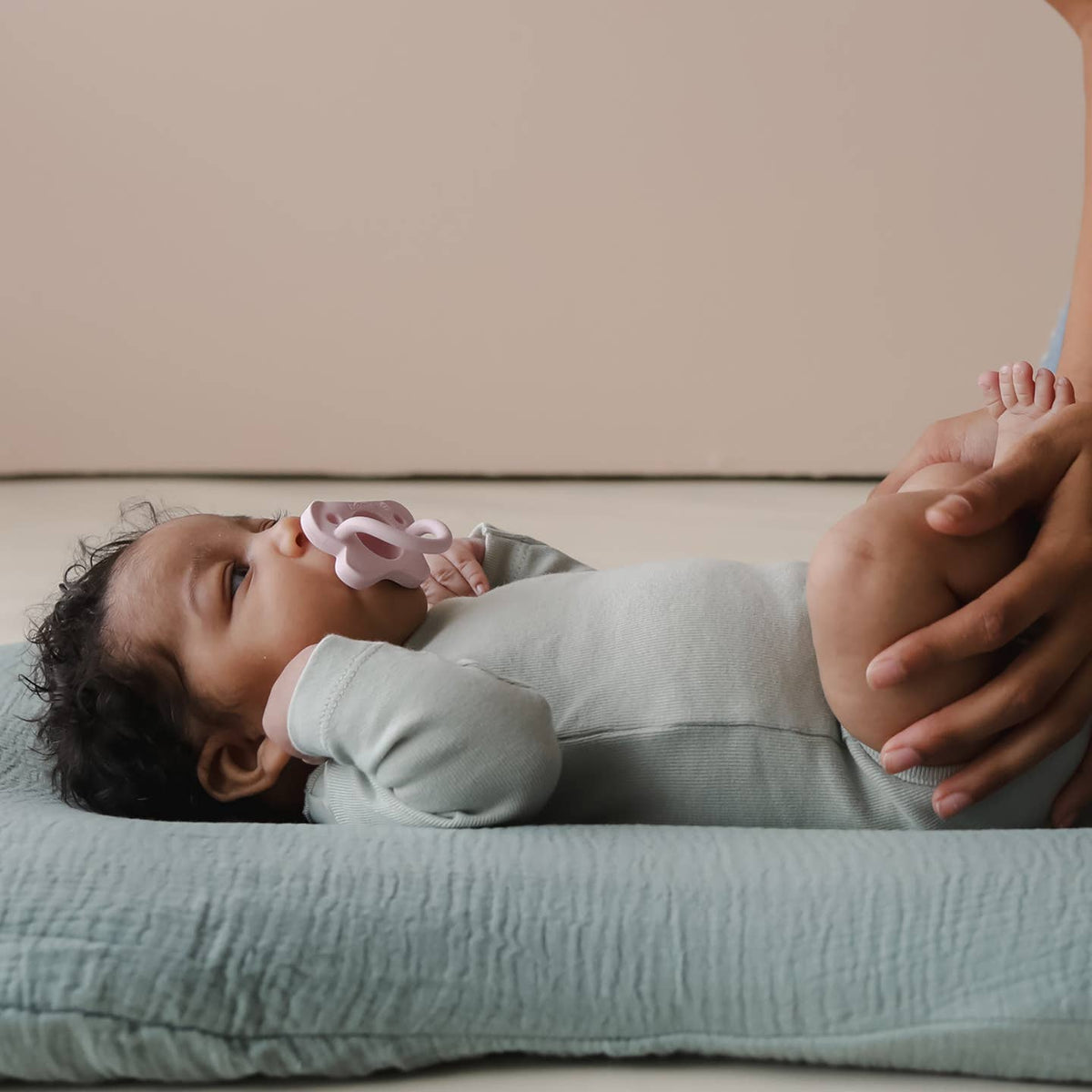 Baby lying on a cushion with a pink teething ring, held by an adult's hand.