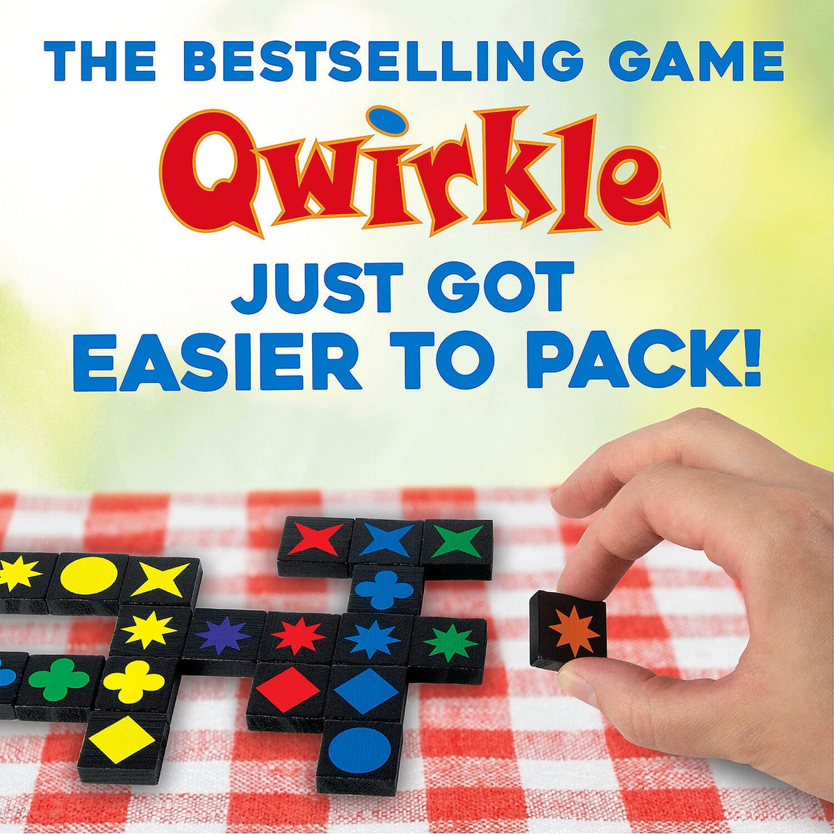 A hand holds a small black game piece with a star symbol over a red-and-white checkered tablecloth, with Qwirkle game boards visible. Large text at the top reads 'THE BESTSELLING GAME Qwirkle JUST GOT EASIER TO PACK!'.