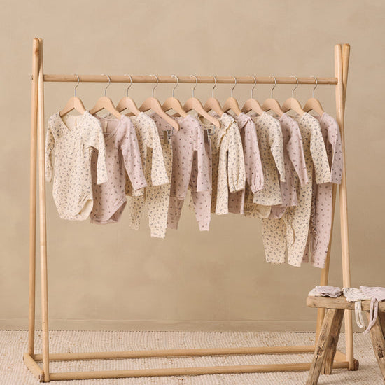 A wooden clothing rack holding multiple baby long-sleeve bodysuits and onesies in soft blush, cream, and beige tones with small polka dots, set against a warm beige wall and textured rug.
