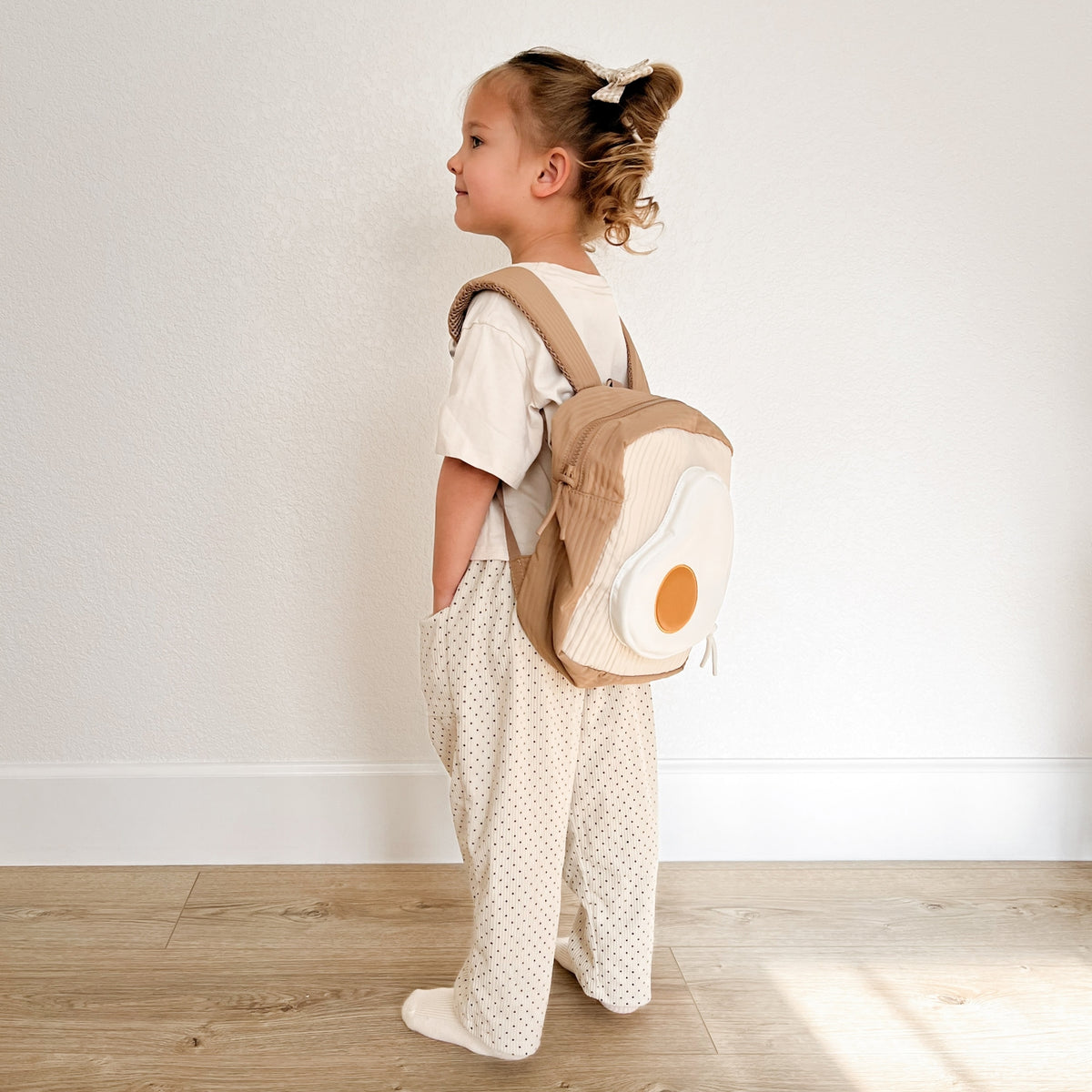 Child wearing a backpack shaped like an egg on a wooden floor.