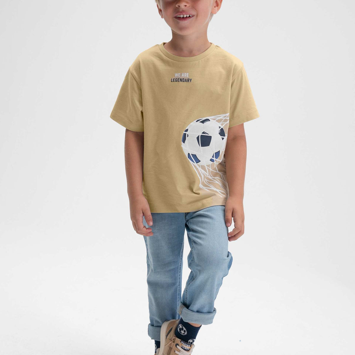 Child wearing a beige t-shirt with a graphic design, light blue jeans, and a cap on a white background