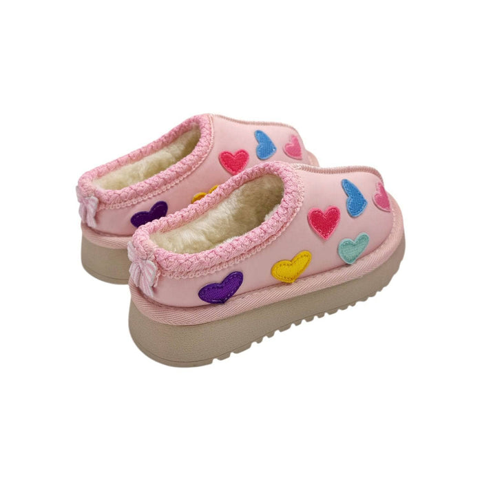 A pair of pink slip-on slippers with a soft cream sherpa lining and colorful embroidered heart patches in pink, blue, yellow, and purple, set against a plain white background.