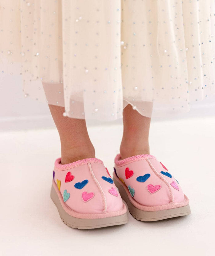 Pink slip-on slippers with colorful heart appliqués, shown on a child's feet with a white tulle skirt visible.