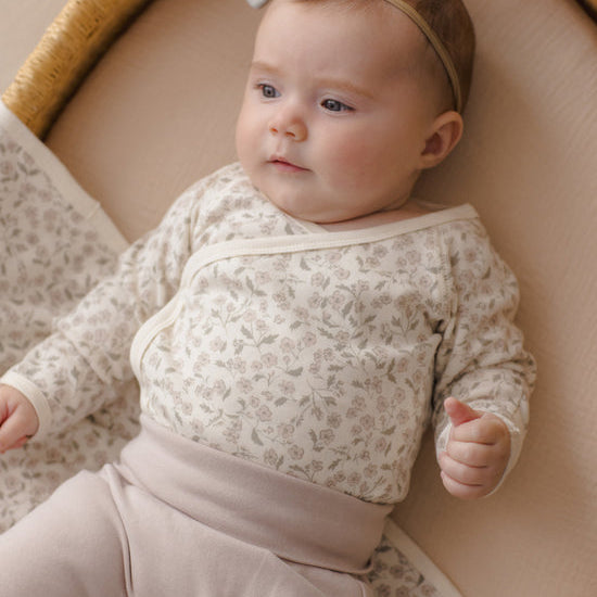A baby lying on a woven bassinet, wearing a cream and beige floral long-sleeve wrap top with matching beige pants and a light gray bow headband.