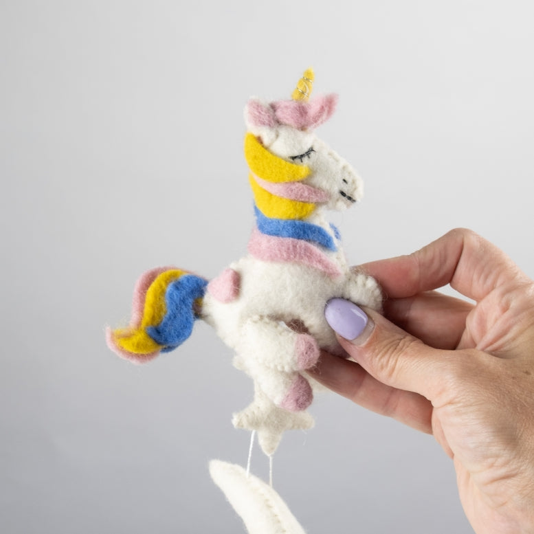 A hand holds a cluster of white felted wool unicorn toys with pastel pink, yellow, and blue accents, attached to a white circular hanging frame against a light gray background.