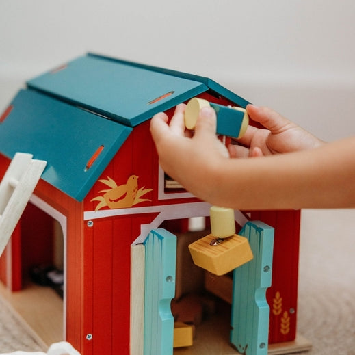 A child’s hands operating the working pulley system on the Wooden Big Red Barn Playset by Mentari, lifting a yellow magnetic hay bale toward the loft while open double doors reveal a glimpse inside the red barn.