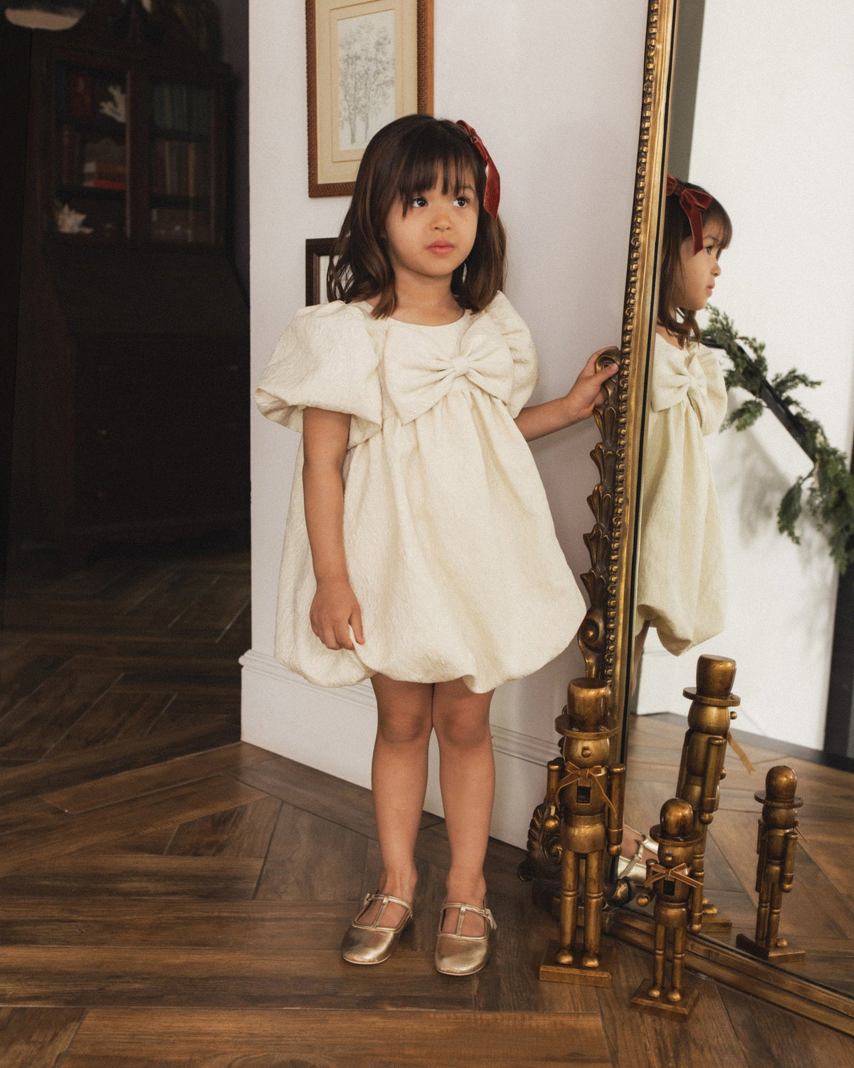 A young girl with dark brown hair wearing a cream-colored dress with a large bow on the chest, standing in a room with a wooden floor and an ornate gold mirror.