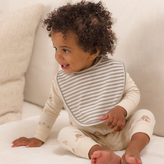 A baby with curly dark hair sits on a white sofa wearing a cream long-sleeve romper with small teddy bear prints and a gray striped bib.