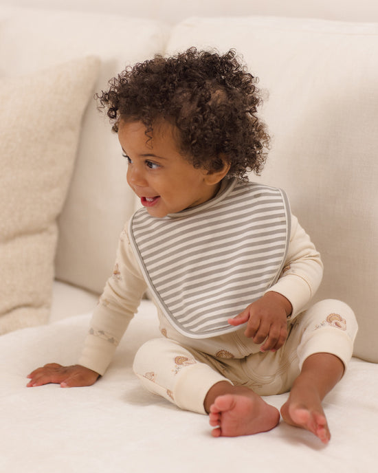 A baby with curly dark hair sits on a white sofa wearing a cream long-sleeve romper with small teddy bear prints and a gray striped bib.