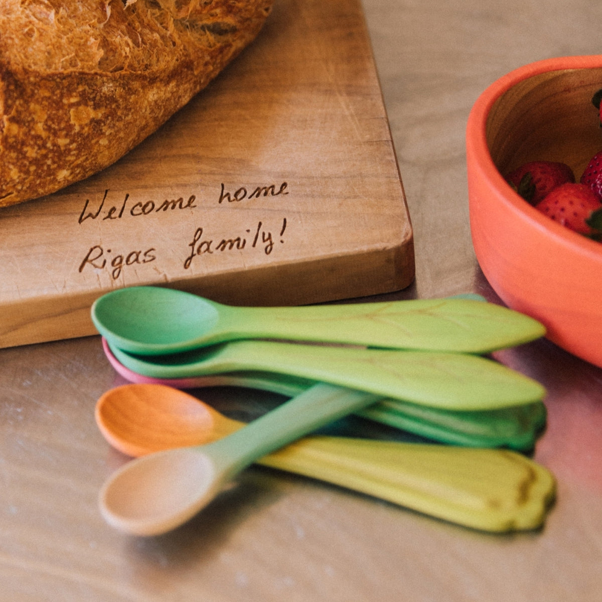 Colorful wooden spoons on a wooden surface with a bowl of strawberries and a cutting board with a message.