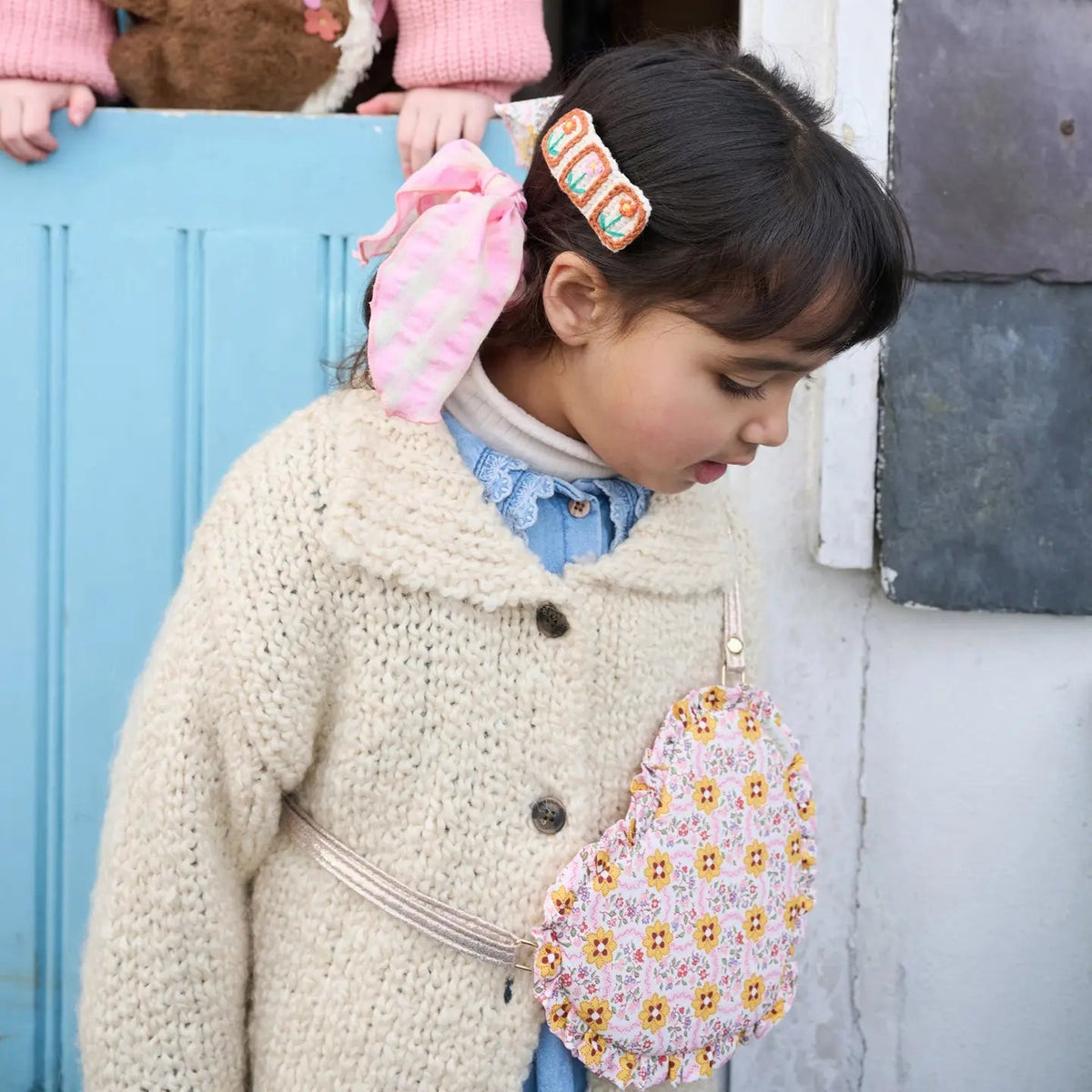 A young girl wearing a chunky cream knit cardigan, blue shirt, and pink hair bow, holding a pink floral heart-shaped purse. She is standing in front of a light blue wooden door.
