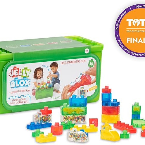The product image displays the Jelly Blox Stash 'n' Stack toy set by Continuum Games, featuring a purple circular badge in the top right corner indicating it as a Toy of the Year (TOTY) Awards 2025 Finalist.
It includes a bright green rectangular storage box with a translucent lid and a product label on the front, alongside several colorful, translucent squishy building blocks and sensory pieces arranged in front of it on a white background.