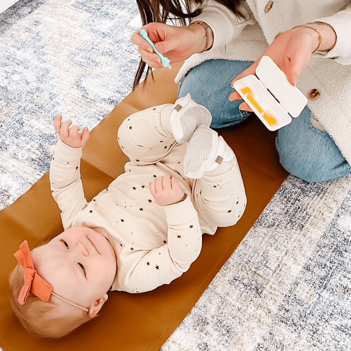 Baby lying on a brown mat with a person holding a pacifier and bottle.