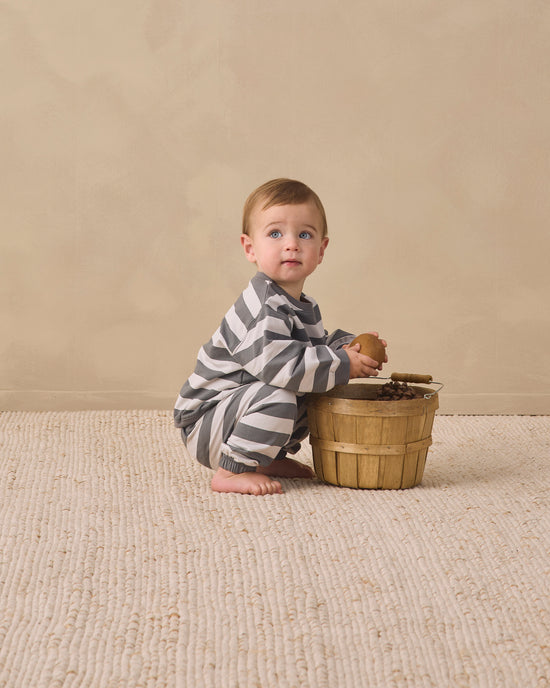 A baby wearing a gray and white striped long-sleeve pajama set sits barefoot on a textured cream rug next to a small wooden basket, looking up and to the side.