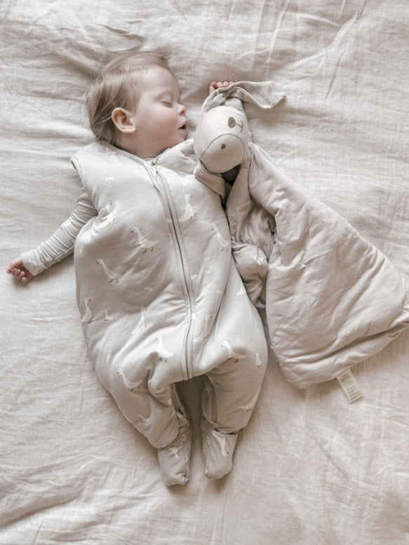 A baby sleeping soundly in a light beige zip-up footed sleeper, lying on a textured cream linen sheet and holding a plush donkey toy.