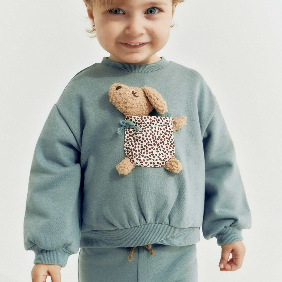 Child wearing a light blue sweatshirt with a teddy bear design on a plain background
