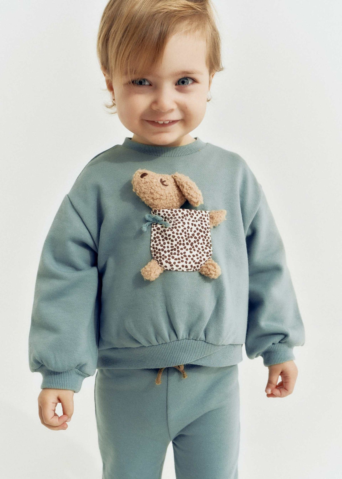 Child wearing a light blue sweatshirt with a teddy bear design on a plain background