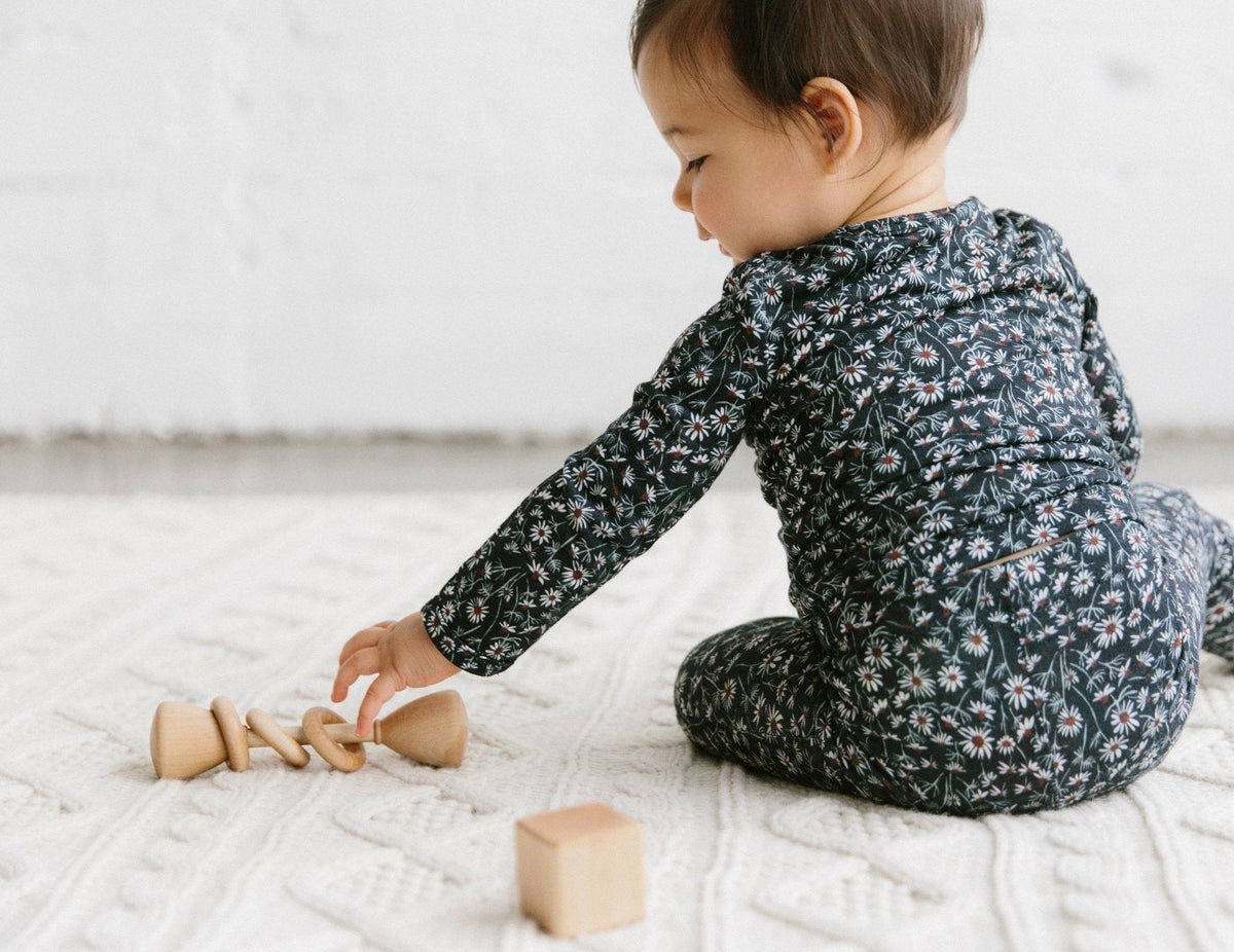 Baby in a floral onesie playing with wooden toys on a white blanket.