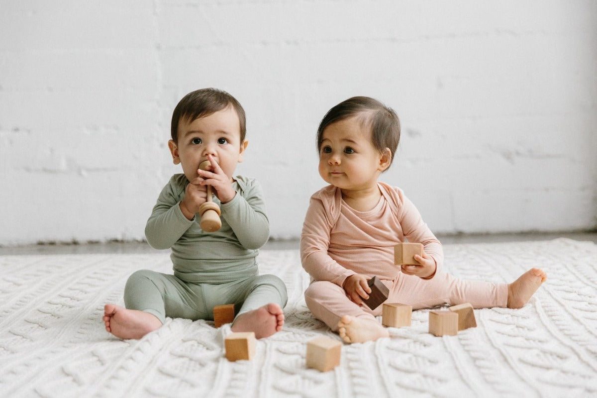 Two babies playing with wooden blocks on a white blanket.