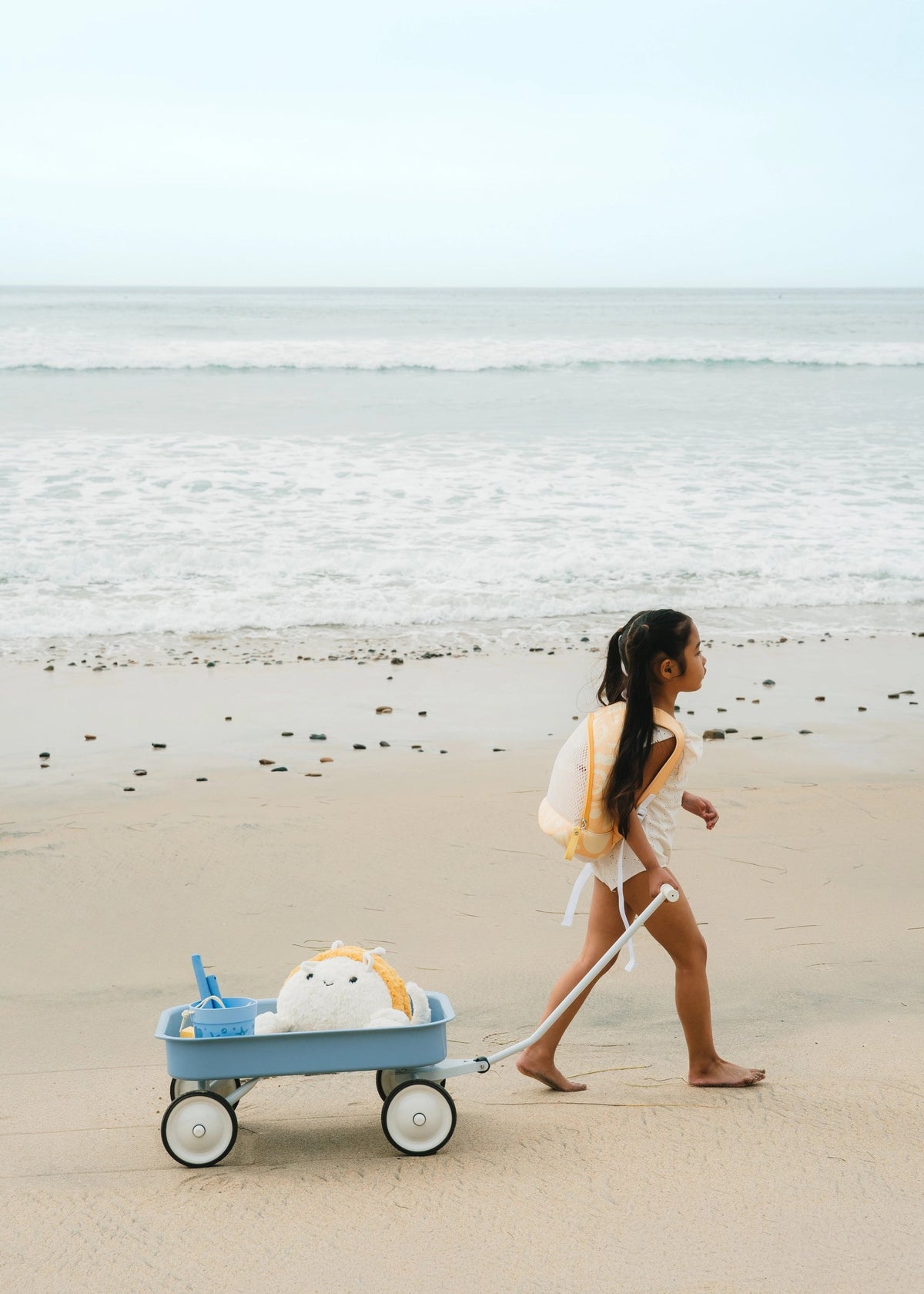 Woman walking on a beach with a blue cart pulling a plush toy