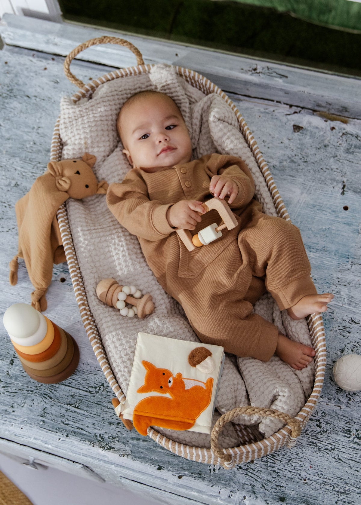 Baby in a brown outfit lying in a basket with toys on a wooden floor.