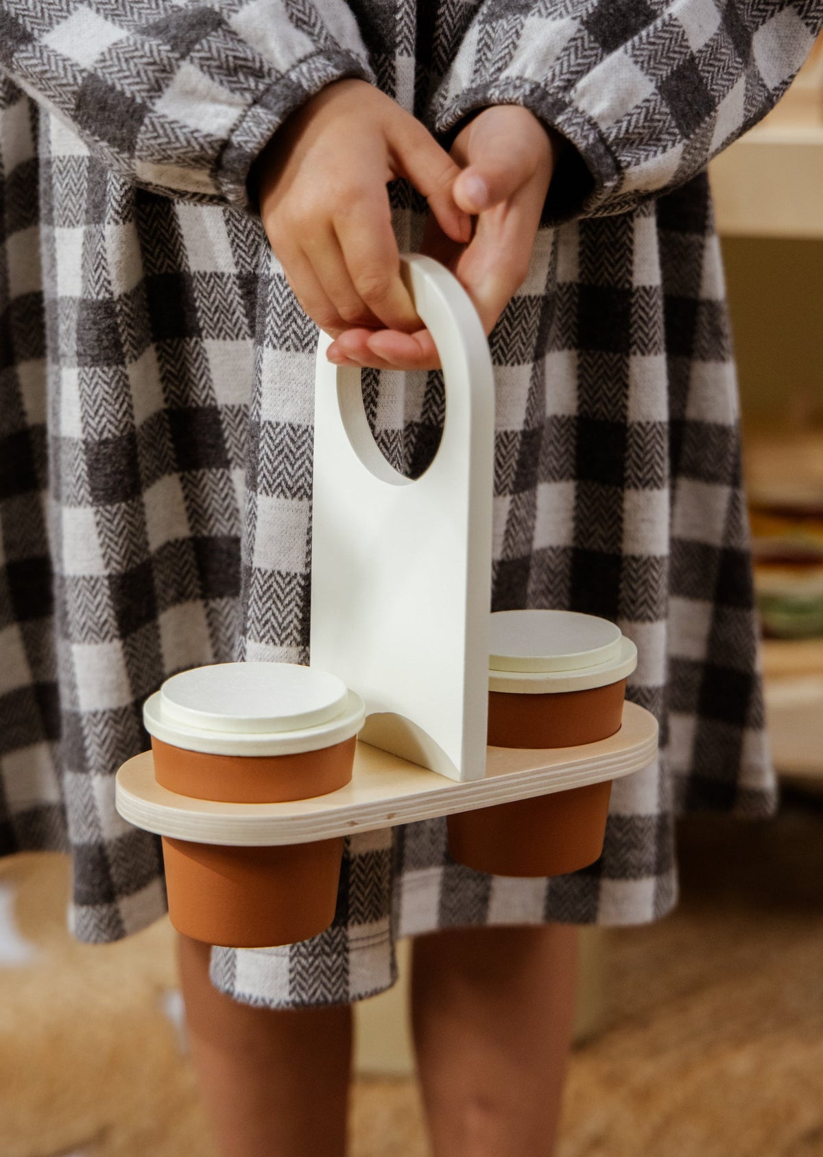 Person holding a wooden cup holder with three brown cups, wearing a checkered robe.