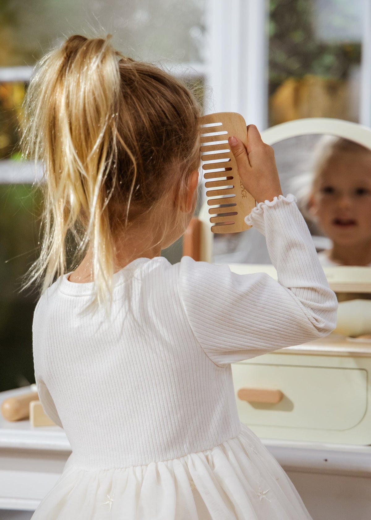 Child in a white dress combing their hair in front of a mirror.