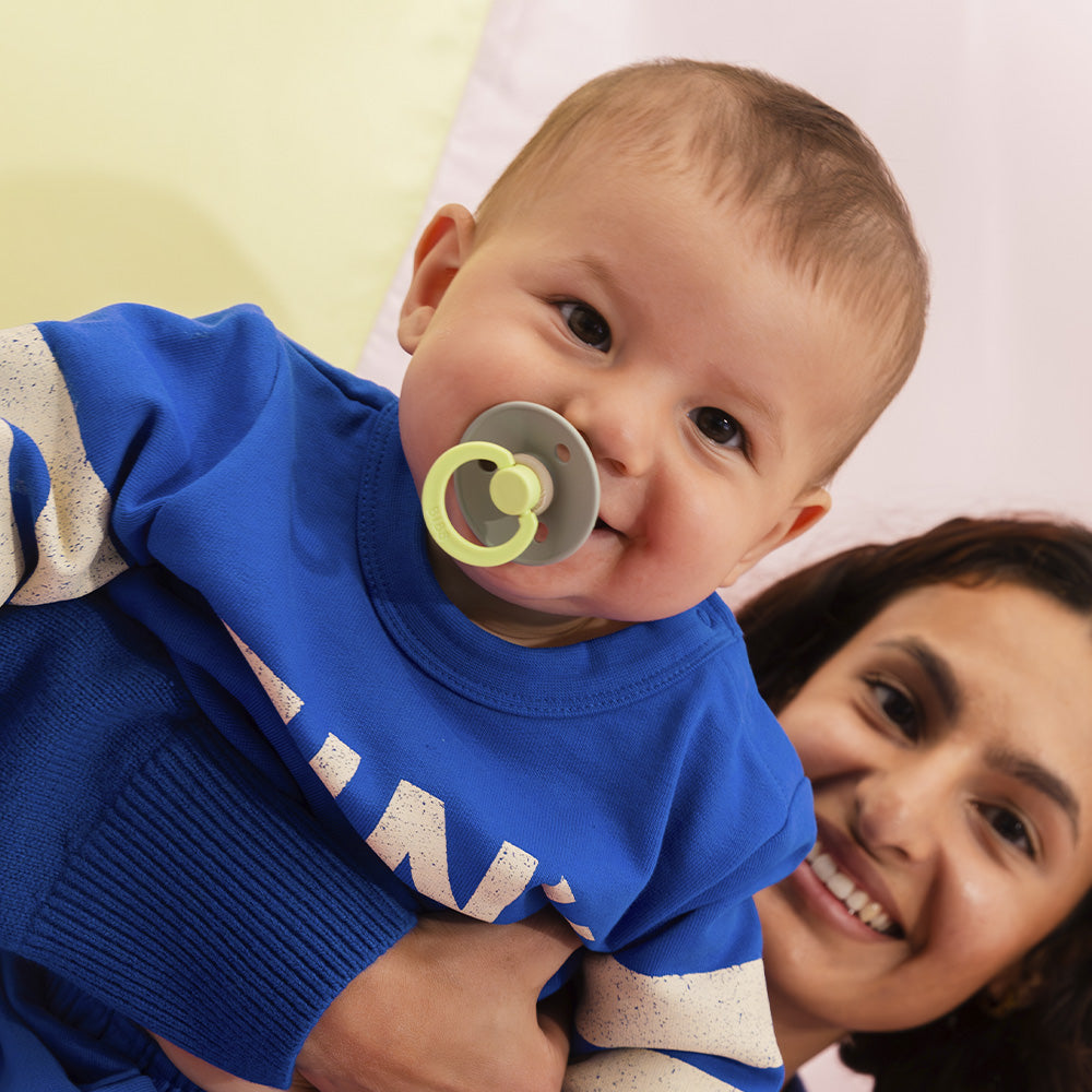 Baby wearing a blue outfit with a pacifier, held by a smiling woman.