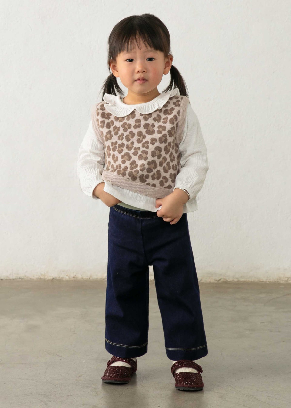 A toddler girl stands against a plain off-white wall wearing a white long-sleeve top with a brown leopard-print vest, dark navy wide-leg pants, and dark red shoes.