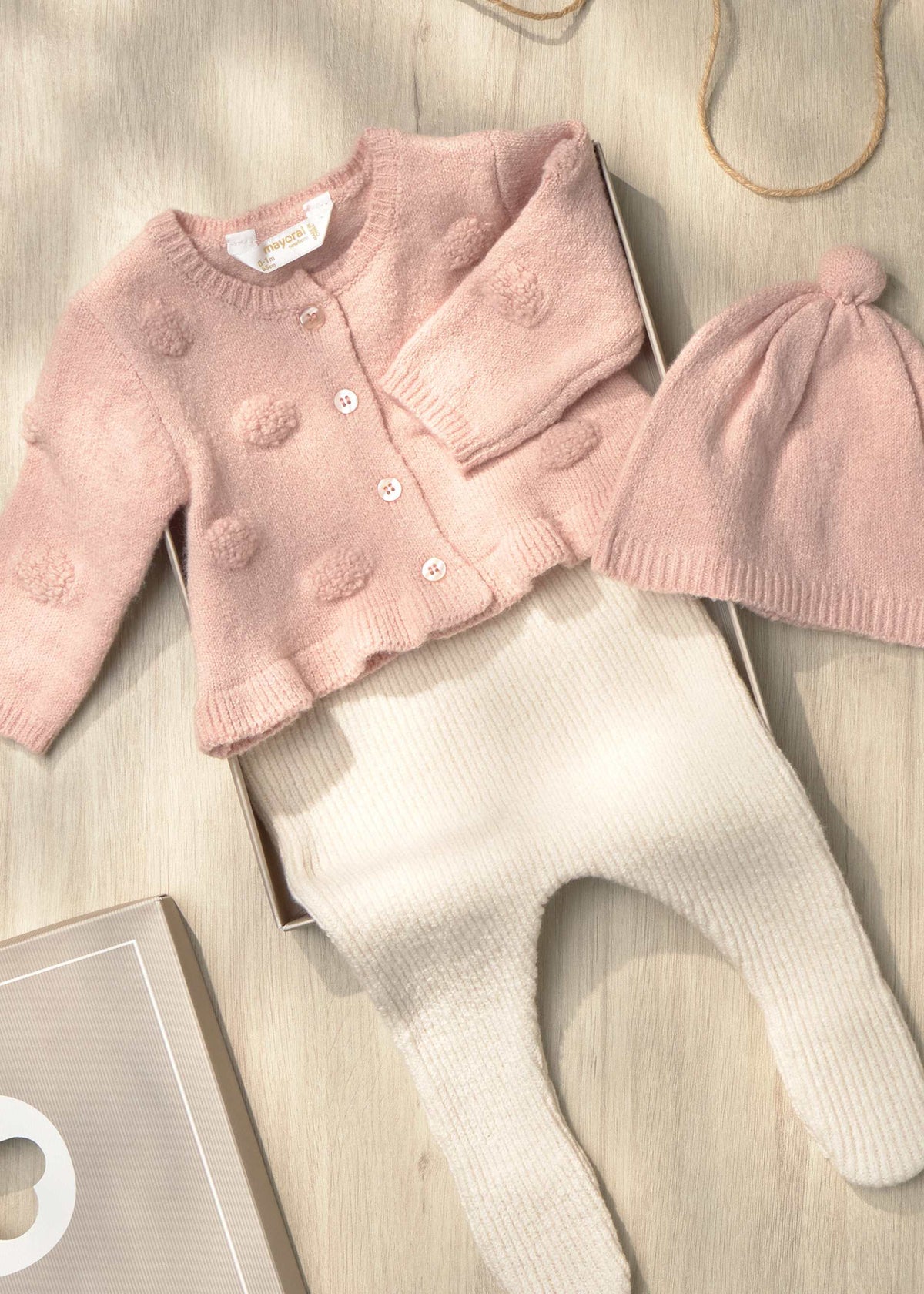 A baby clothing set laid flat on a light wood surface, including a pale pink knitted cardigan with pom-pom texture, cream ribbed footed pants, and a matching pink knit hat with a pom-pom. A small box is partially visible underneath.