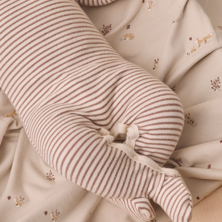 A close-up of a long, cylindrical nursing pillow with thin brown and beige stripes, nestled in a beige patterned blanket with small embroidered deer.