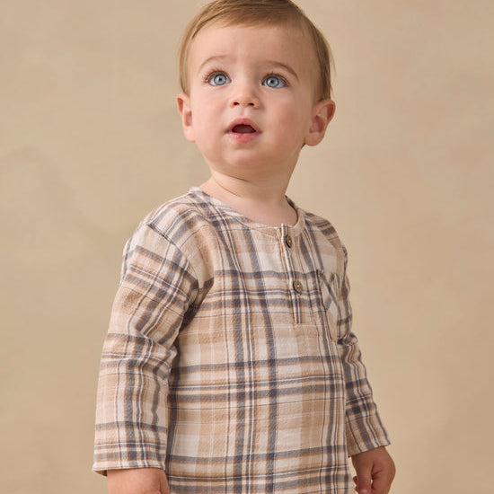 A baby boy wearing a beige and brown plaid long-sleeve henley shirt, standing against a solid beige studio background.