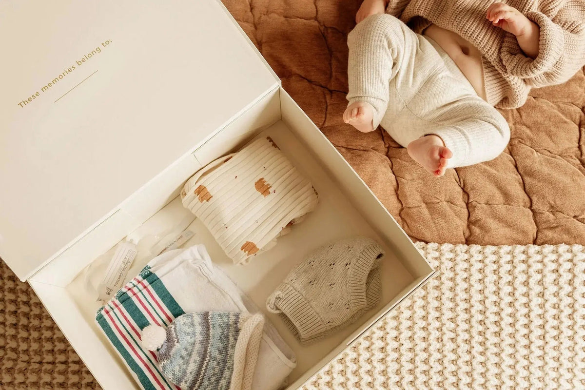 Baby outfit set in a box with a baby lying on a quilted surface