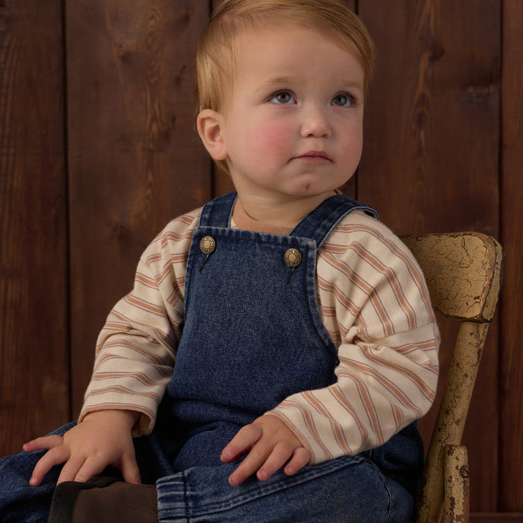 A baby boy with light hair sits on a rustic wooden chair against a dark wood plank background, wearing a beige and brown striped long-sleeve shirt under blue denim overalls.