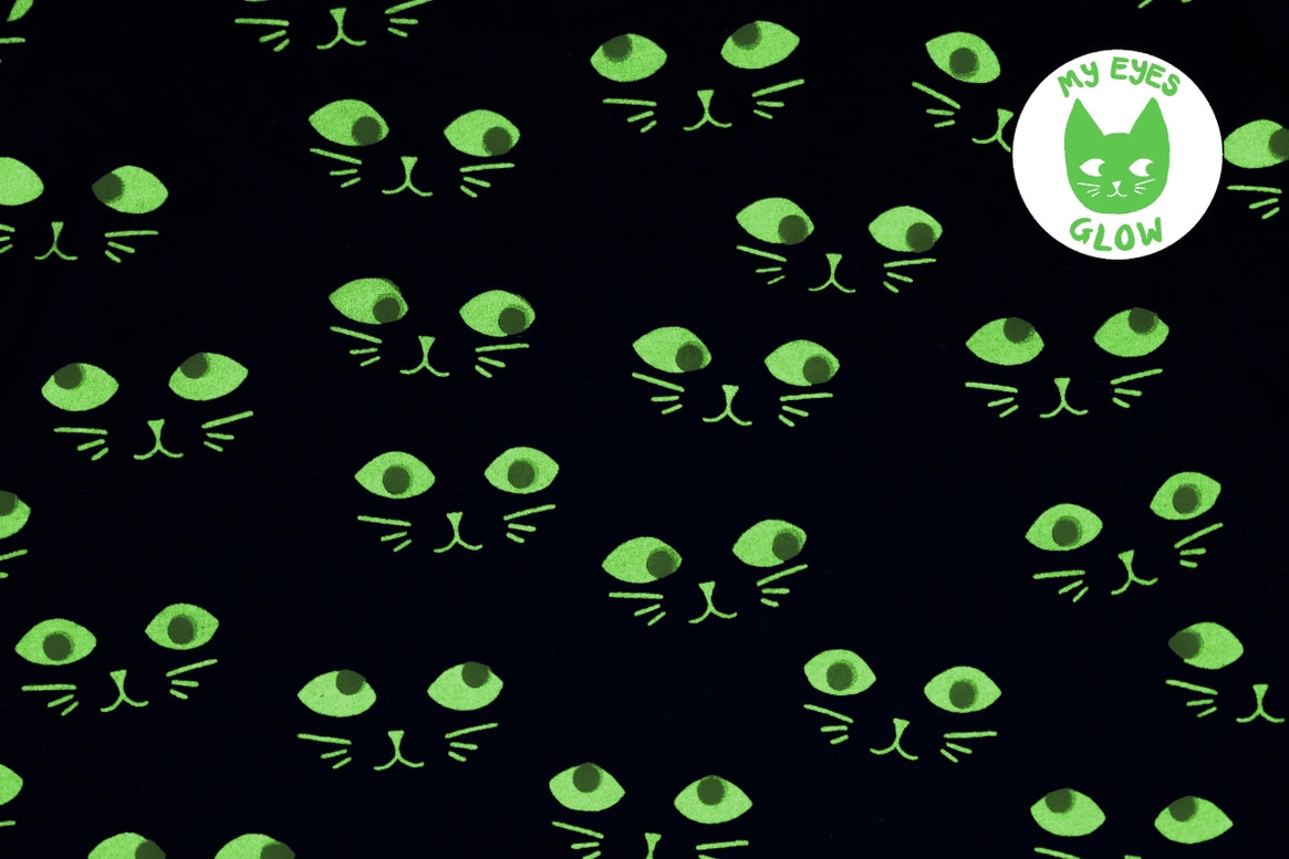 Black fabric with a repeating pattern of glowing neon green cat faces, featuring a circular 'My Eyes Glow' logo in the top right corner.