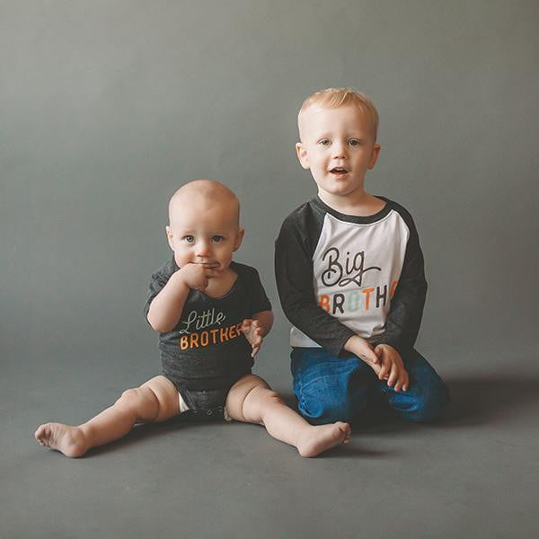 Two young boys wearing 'Little Brother' and 'Big Brother' shirts on a gray background