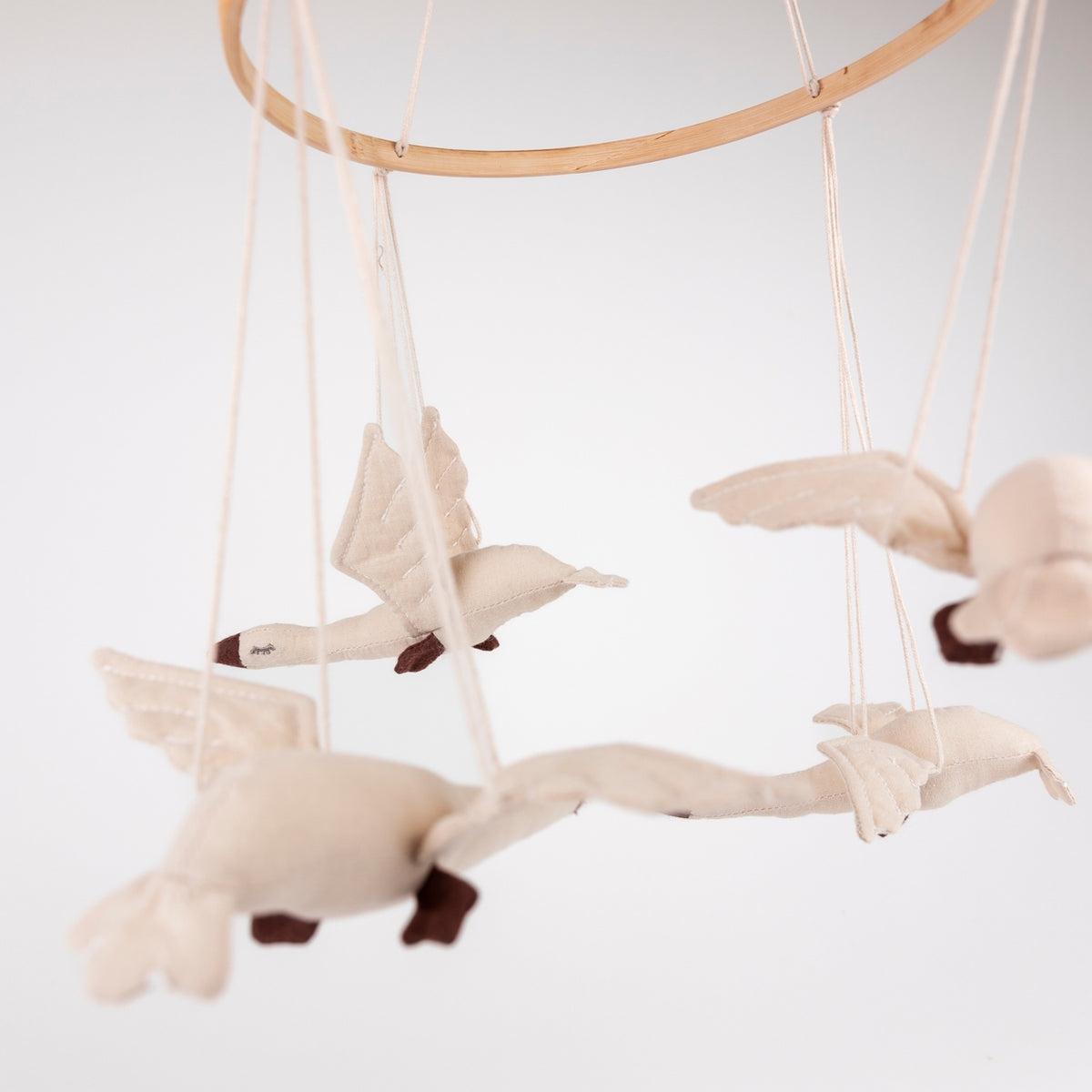 A fabric baby mobile featuring several cream-colored goose figures with dark brown beaks and legs, suspended from a natural wood hoop against a white background.